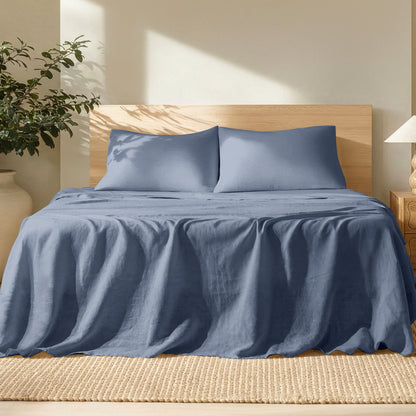Bed with linen bedding and pillows in a bright room with a plant and lamp.