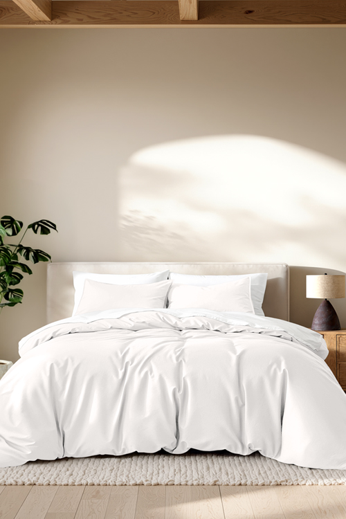 A neatly made bed with white bedding and pillows in a softly lit bedroom, with a wooden nightstand, lamp, and warm beige wall in the background.