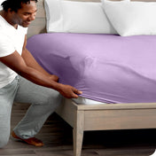 Man in white shirt and gray pants adjusting a fitted sheet on a bed.