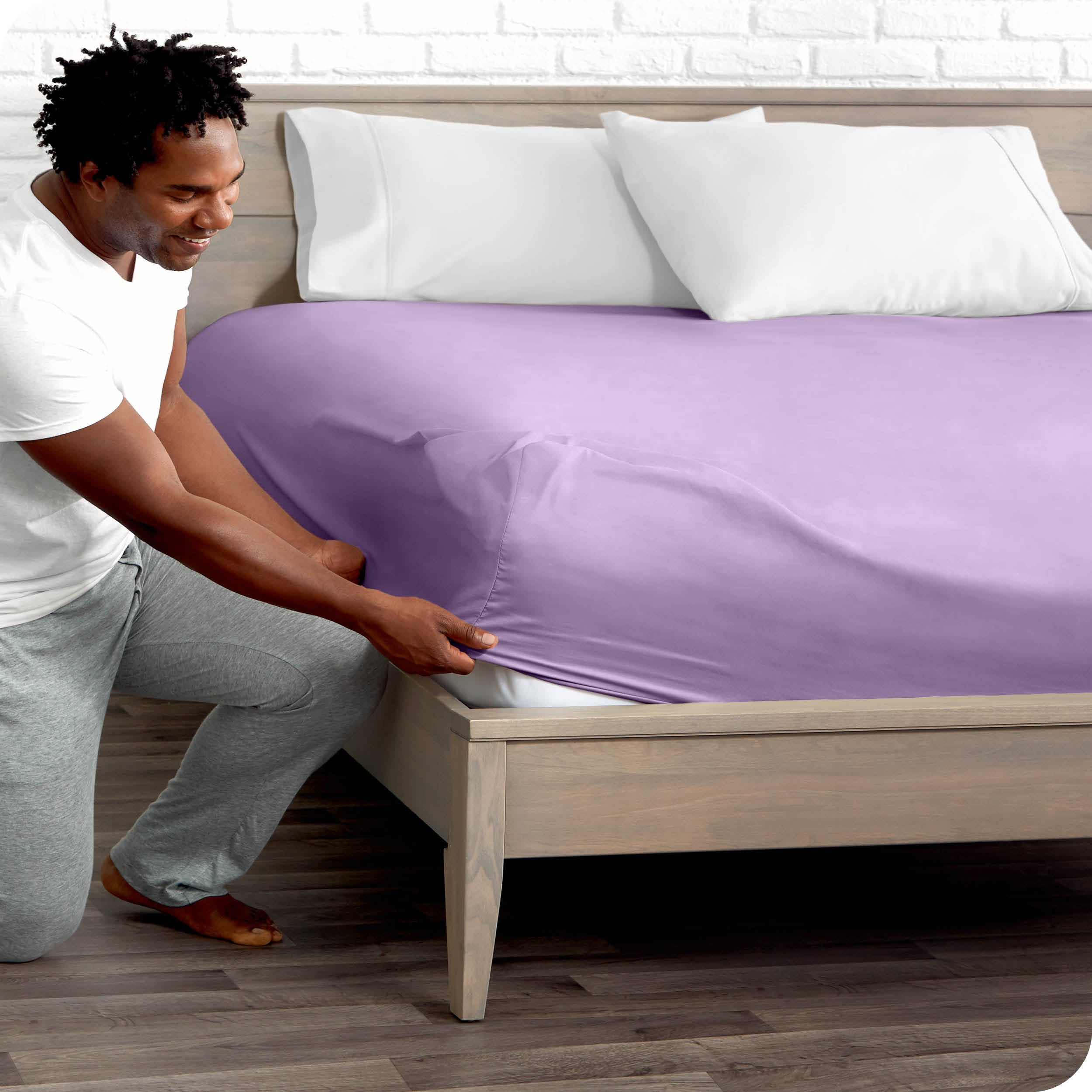 A man is kneeing and putting the corner of the fitted sheet on the mattress. The mattress is on a wooden bed frame. 