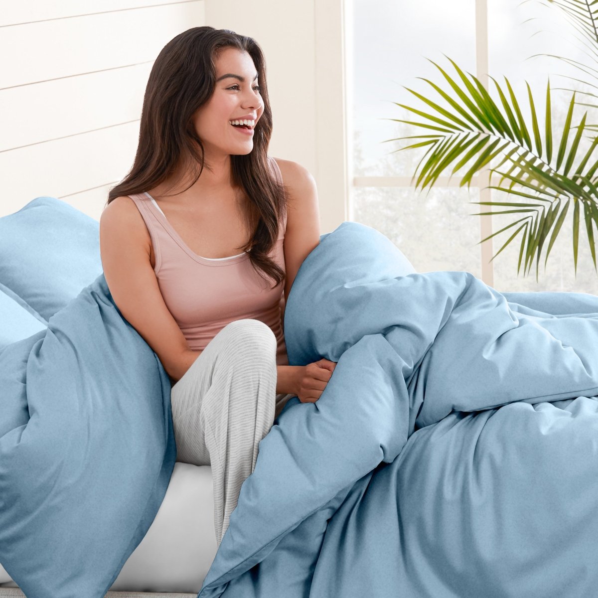 Woman sitting on a bed under duvet cover with a plant in the background