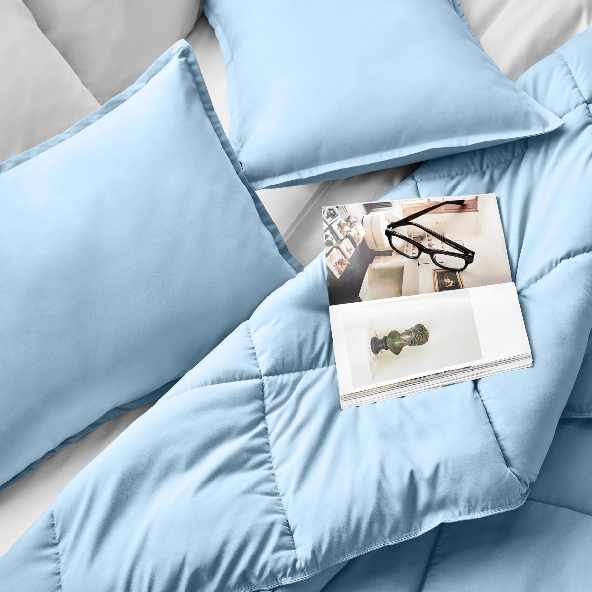 Comforter and pillows on a bed with a magazine and glasses.