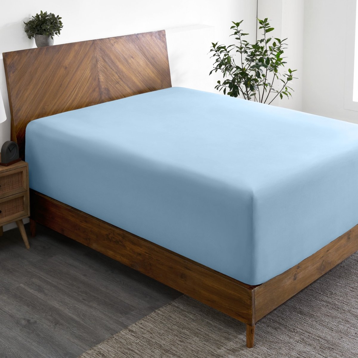 Wooden bed frame with a fitted sheet on the mattress in a room with plants and a nightstand.