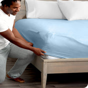 Man in white shirt and gray pants adjusting a fitted sheet on a bed.