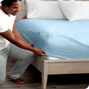 A man is kneeling while putting a fitted sheet on a bed