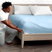 A man is kneeing and putting the corner of the fitted sheet on the mattress. The mattress is on a wooden bed frame.