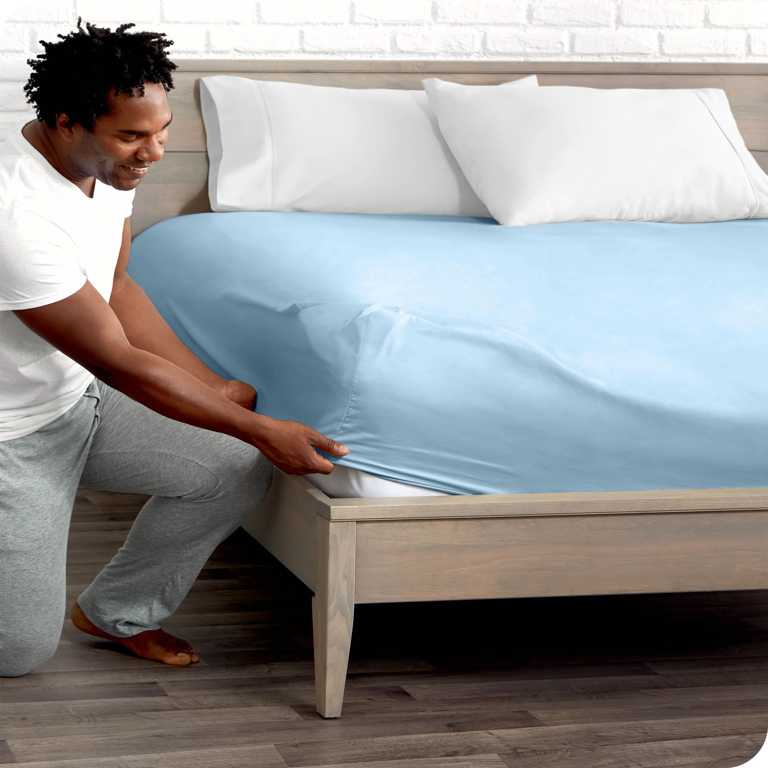 A man is kneeing and putting the corner of the fitted sheet on the mattress. The mattress is on a wooden bed frame.