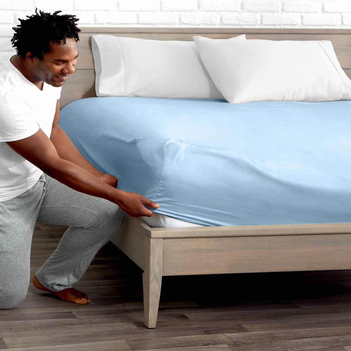 Man adjusting fitted sheet on a bed with white pillows.