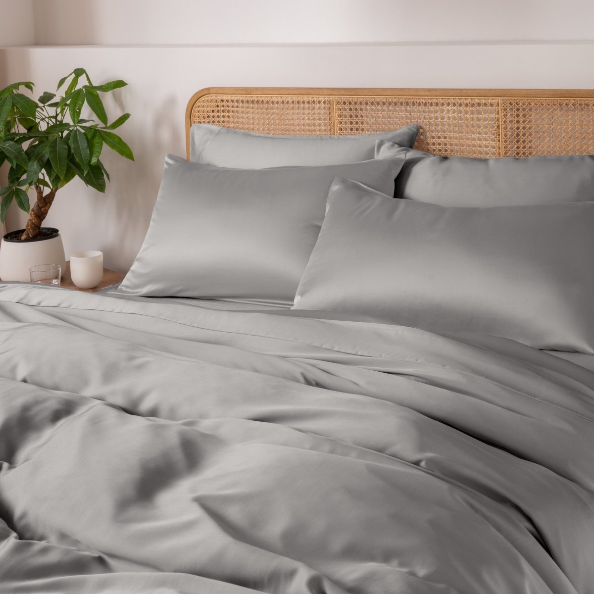 Light grey bedding set on a bed with a wicker headboard and plant in the corner.