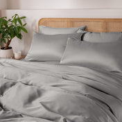 Light grey bedding set on a bed with a wicker headboard and plant in the corner.