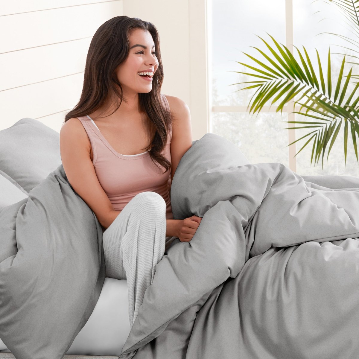 Woman sitting on a bed under duvet cover with a plant in the background