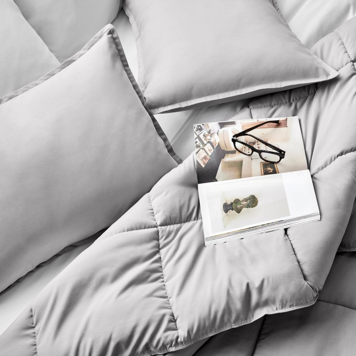 Comforter and pillows on a bed with a magazine and glasses.