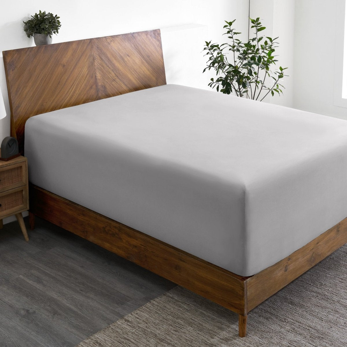 Wooden bed frame with a fitted sheet on the mattress in a room with plants and a nightstand.