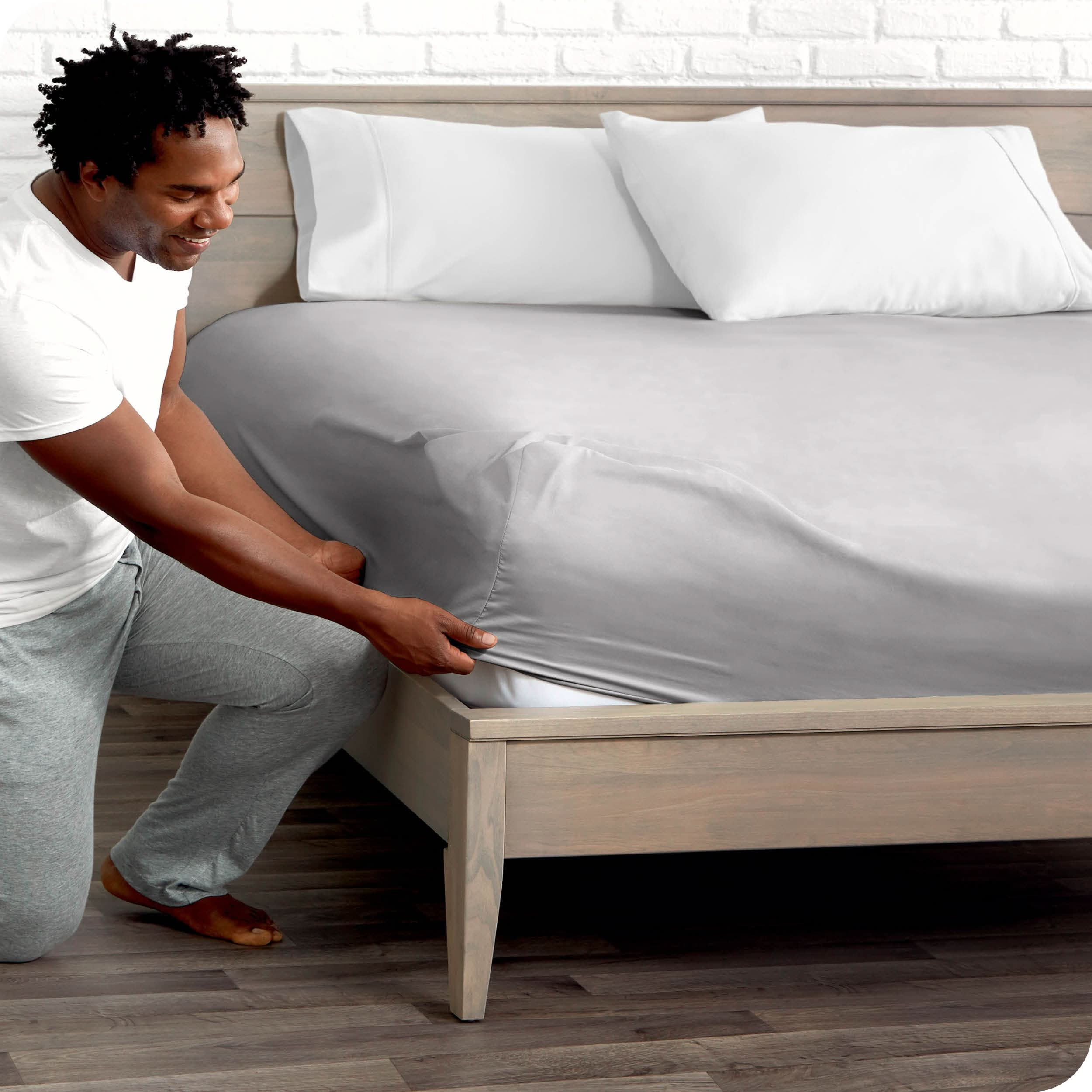 A man is kneeing and putting the corner of the fitted sheet on the mattress. The mattress is on a wooden bed frame.