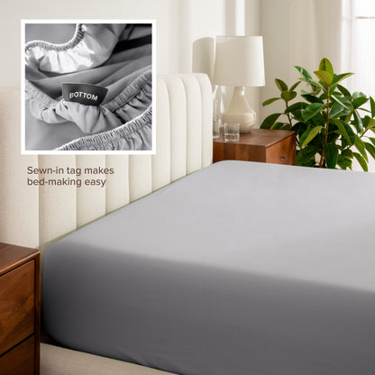 Fitted sheet on a bed with a close-up of a sewn-in tag, nightstand, lamp, and plant in a bedroom setting.