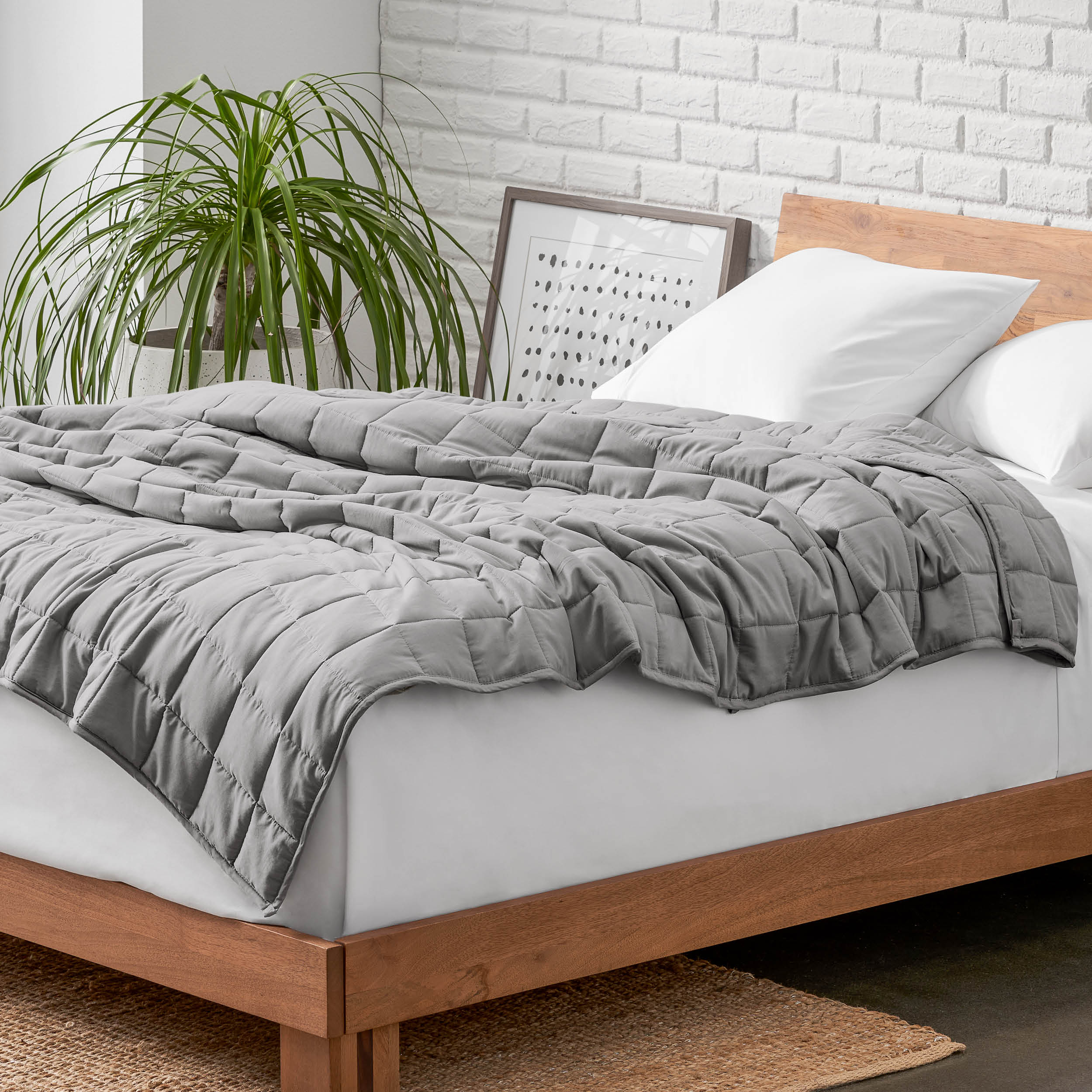 Weighted blanket on a bed with white pillows and a wooden headboard in a room with a plant and brick wall.