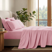 A neatly made bed with in a softly lit bedroom. A large green plant stands beside the bed, and a window with curtains is visible in the background.