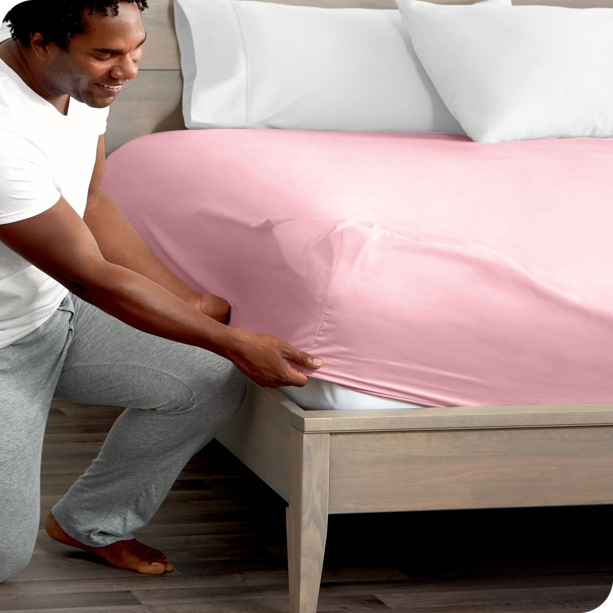 Man in white shirt and gray pants adjusting a fitted sheet on a bed.