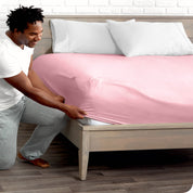 A man is kneeing and putting the corner of the fitted sheet on the mattress. The mattress is on a wooden bed frame. 