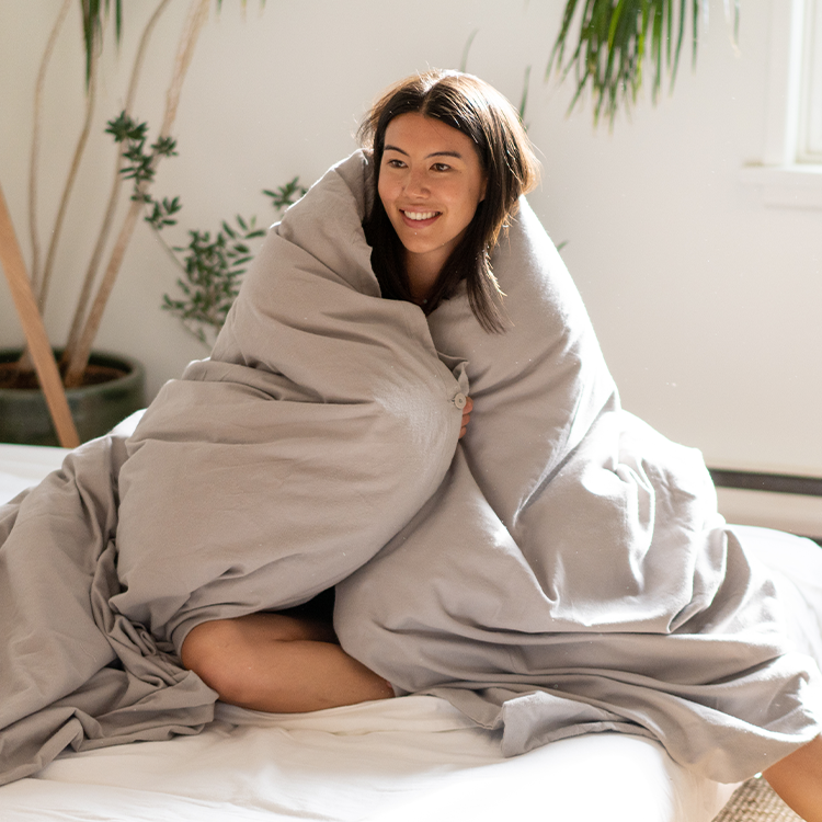 Woman wrapped in a large gray blanket in a cozy room with plants.