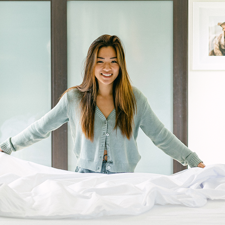 Woman in a light gray cardigan standing behind a bed with white bedding.