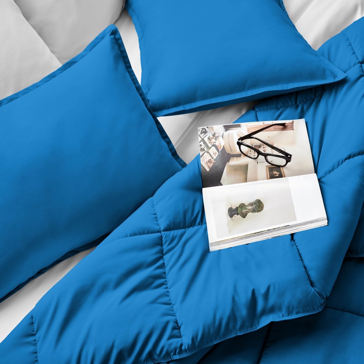 Comforter and pillows on a bed with a magazine and glasses.