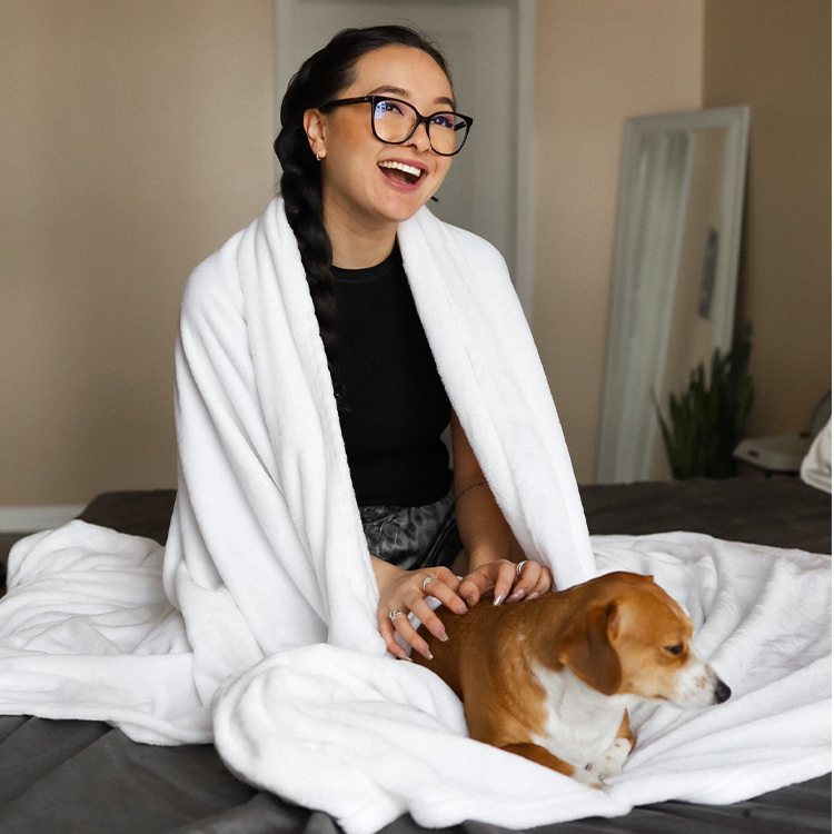 Woman sitting on a bed with a dog, wrapped in a white blanket, in a home setting.