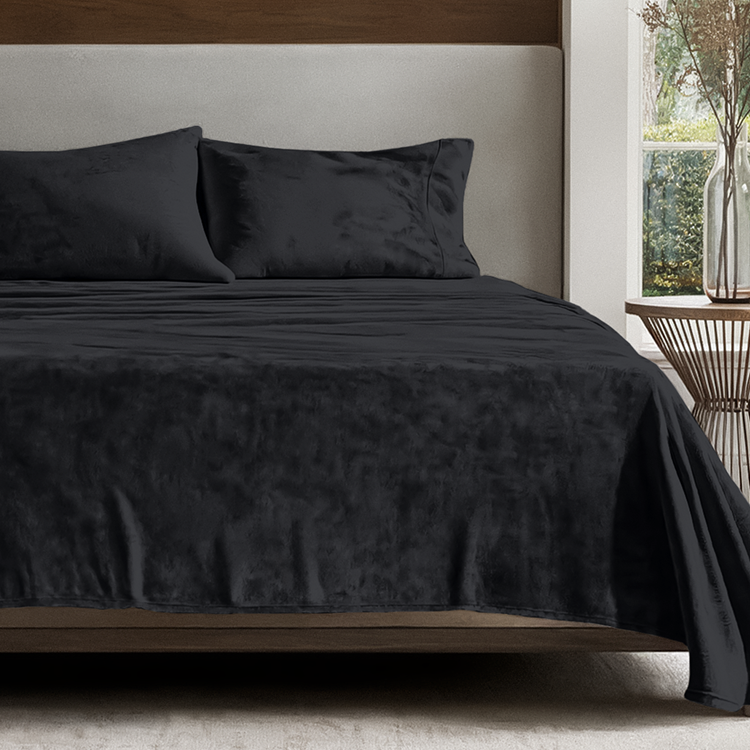 Bed with dark gray bedding and pillows in a room with a window and plant.
