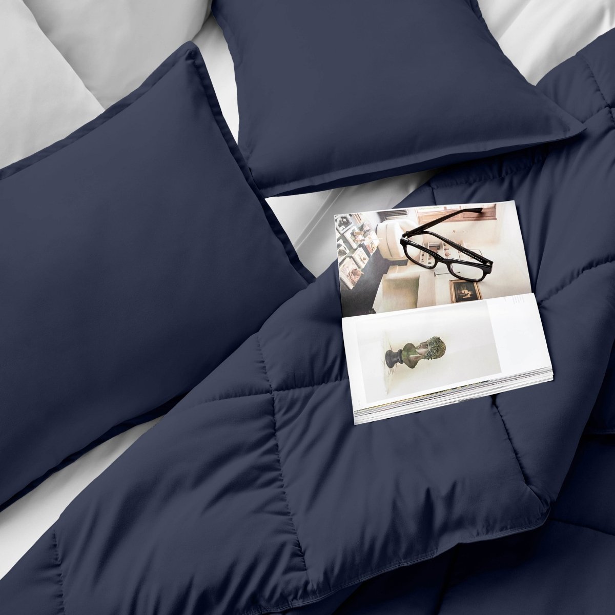Comforter and pillows on a bed with a magazine and glasses.