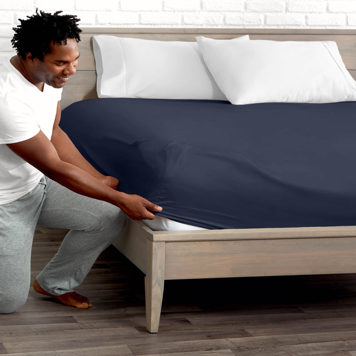 Man adjusting fitted sheet on a bed with white pillows.
