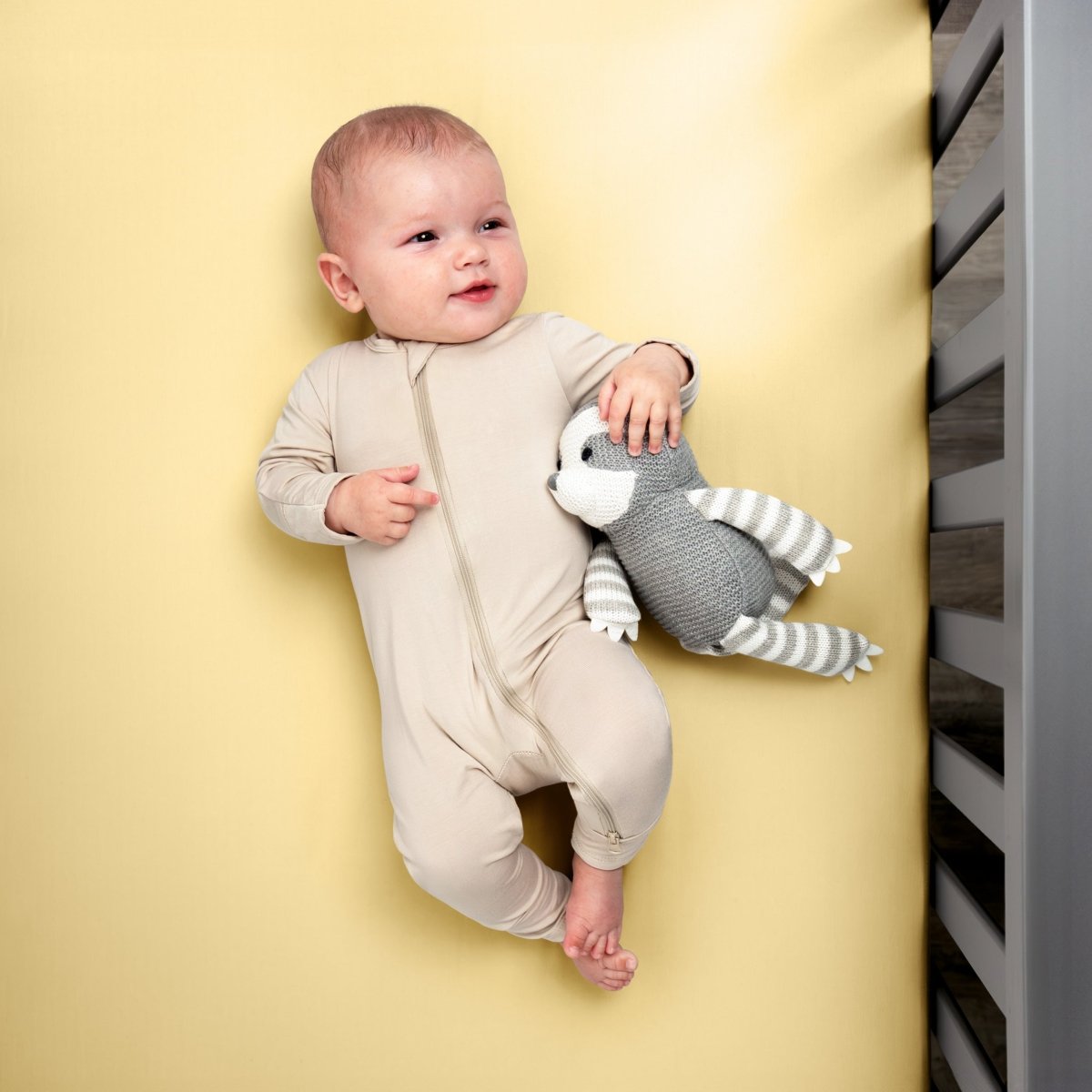 Baby in a beige onesie holding a gray and white striped toy while in a crib