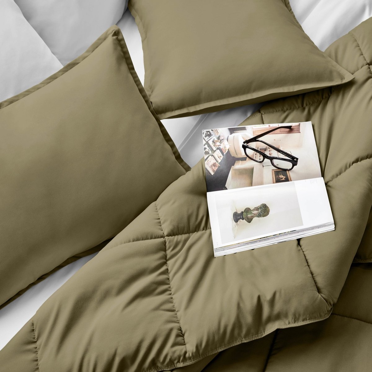Comforter and pillows on a bed with a magazine and glasses.