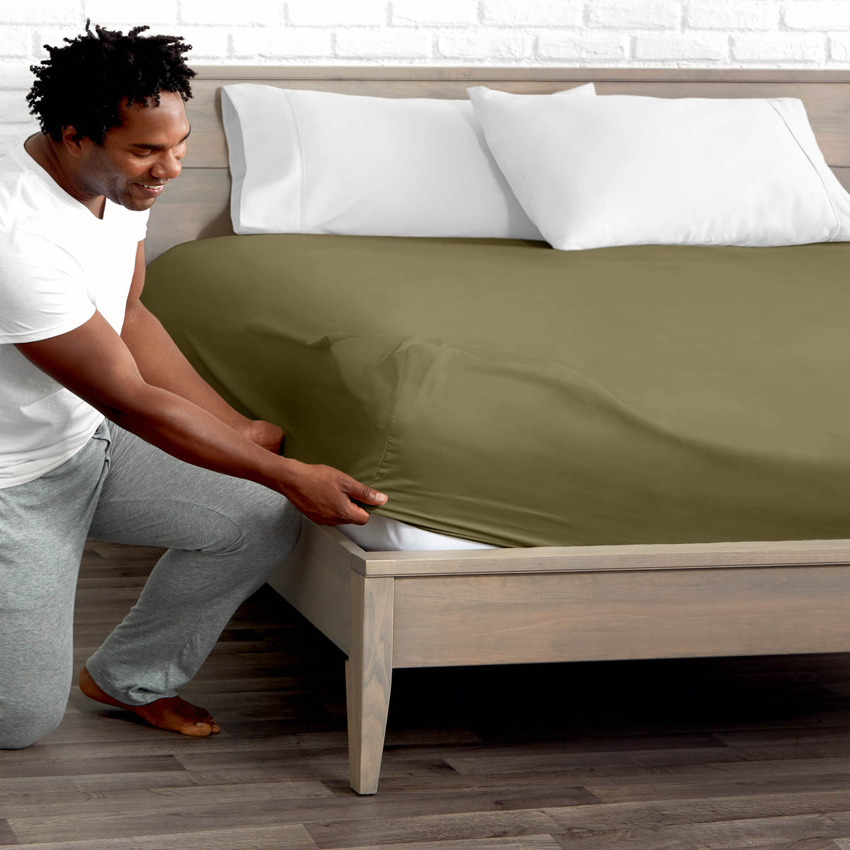 Man adjusting fitted sheet on a bed with white pillows.