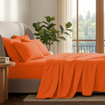 A neatly made bed with in a softly lit bedroom. A large green plant stands beside the bed, and a window with curtains is visible in the background.