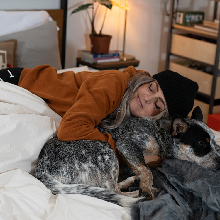 Woman in a brown sweater and black beanie lying on a bed with a dog, surrounded by a cozy bedroom setting.