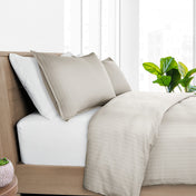 Neatly made bed with beige and white bedding, pillows, and a plant in a bright room.