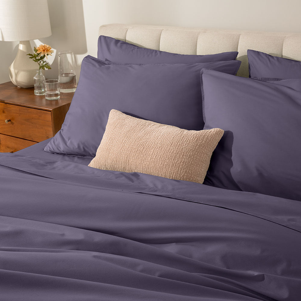 Purple bedding set on a bed with a wooden nightstand and decorative items.