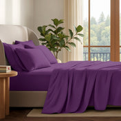A neatly made bed with in a softly lit bedroom. A large green plant stands beside the bed, and a window with curtains is visible in the background.