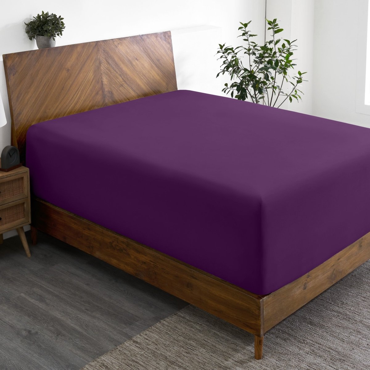 Wooden bed frame with a fitted sheet on the mattress in a room with plants and a nightstand.