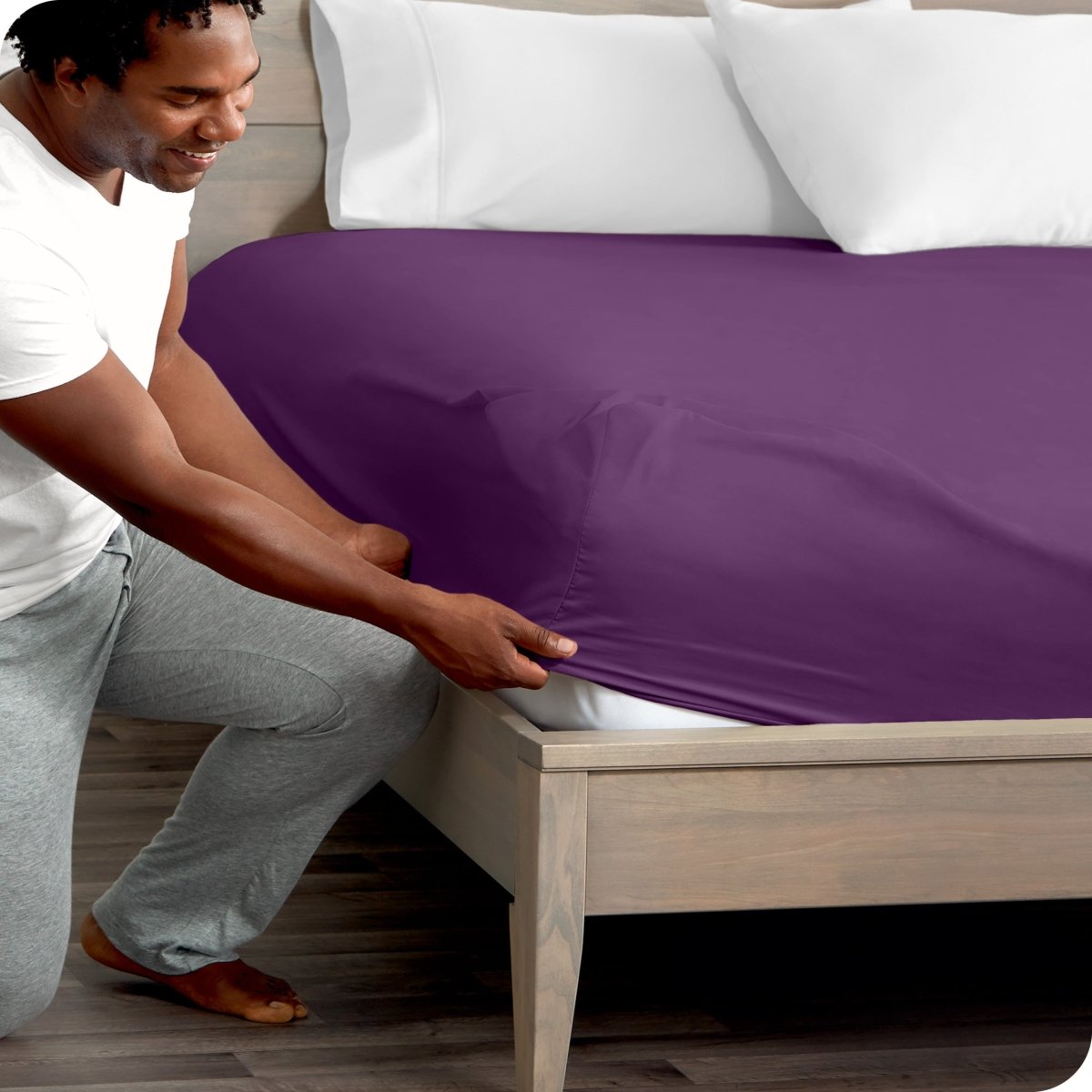 Man in white shirt and gray pants adjusting a fitted sheet on a bed.