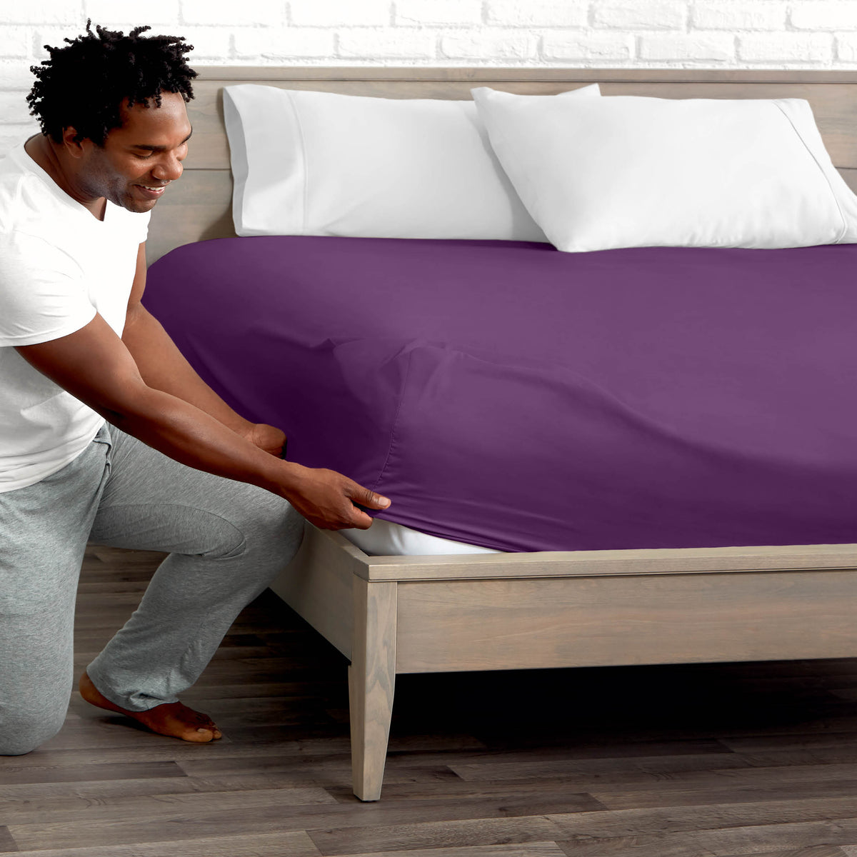 Man adjusting fitted sheet on a bed with white pillows.