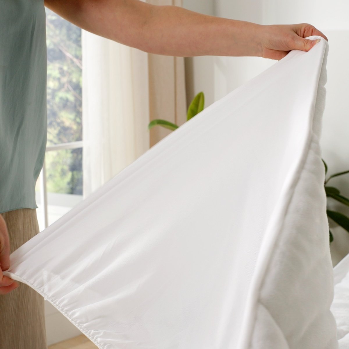 Person holding a white mattress pad over a bed with a blurred window in the background