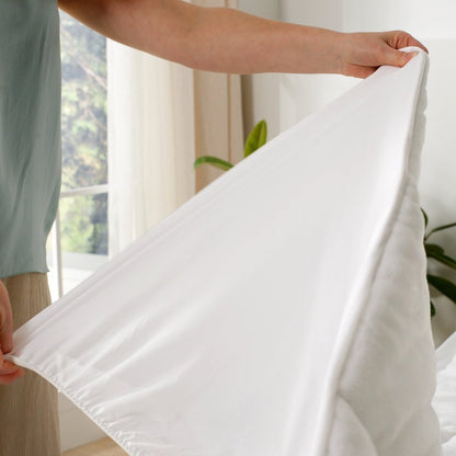 Person holding a white mattress pad over a bed with a blurred window in the background