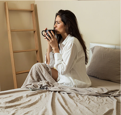 Woman in a white robe sitting on a bed with tan sheets holding a coffee mug