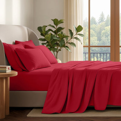 A neatly made bed with in a softly lit bedroom. A large green plant stands beside the bed, and a window with curtains is visible in the background.
