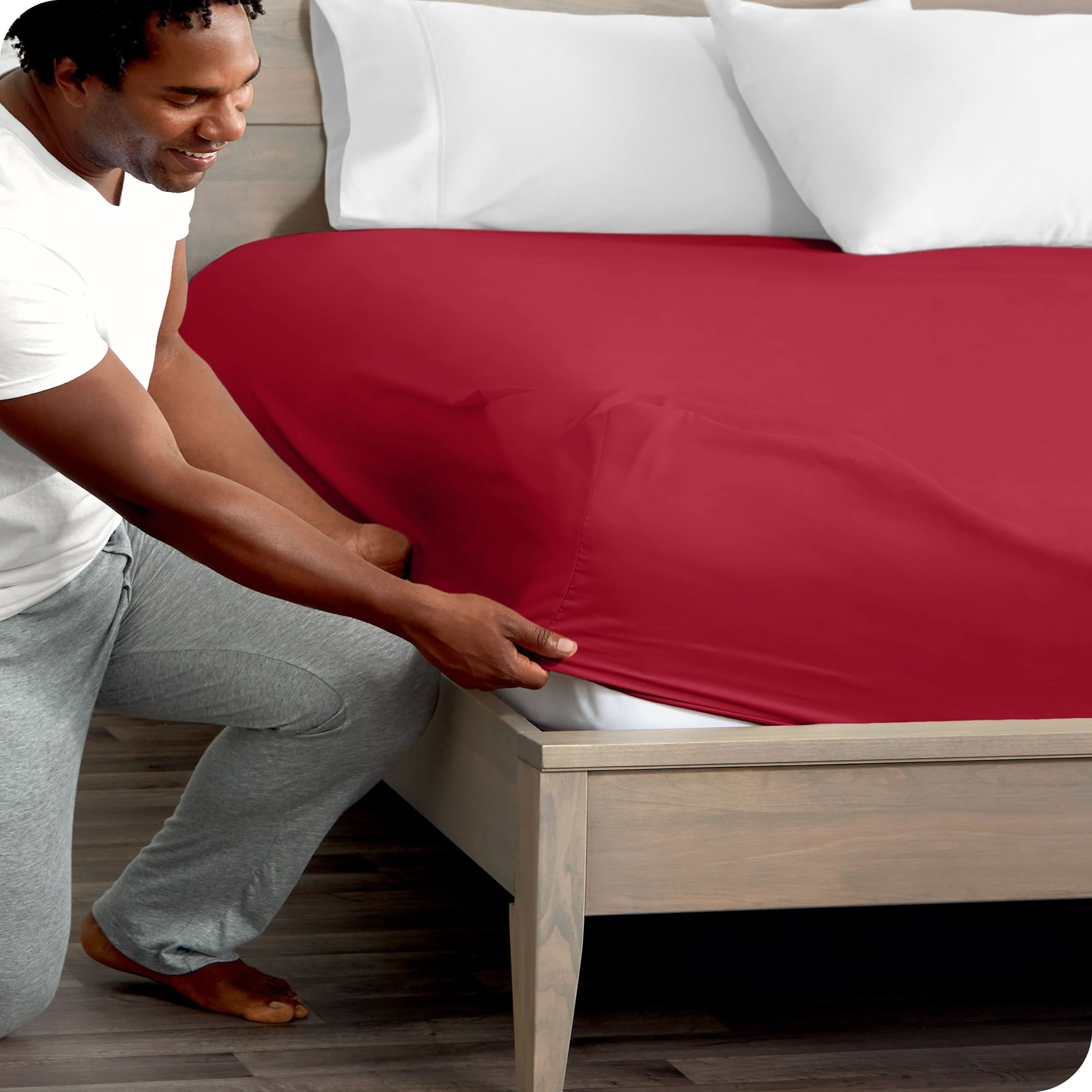 A man is kneeling while putting a fitted sheet on a bed