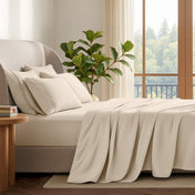 A neatly made bed with in a softly lit bedroom. A large green plant stands beside the bed, and a window with curtains is visible in the background.