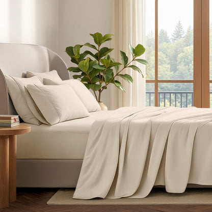 A neatly made bed with in a softly lit bedroom. A large green plant stands beside the bed, and a window with curtains is visible in the background.