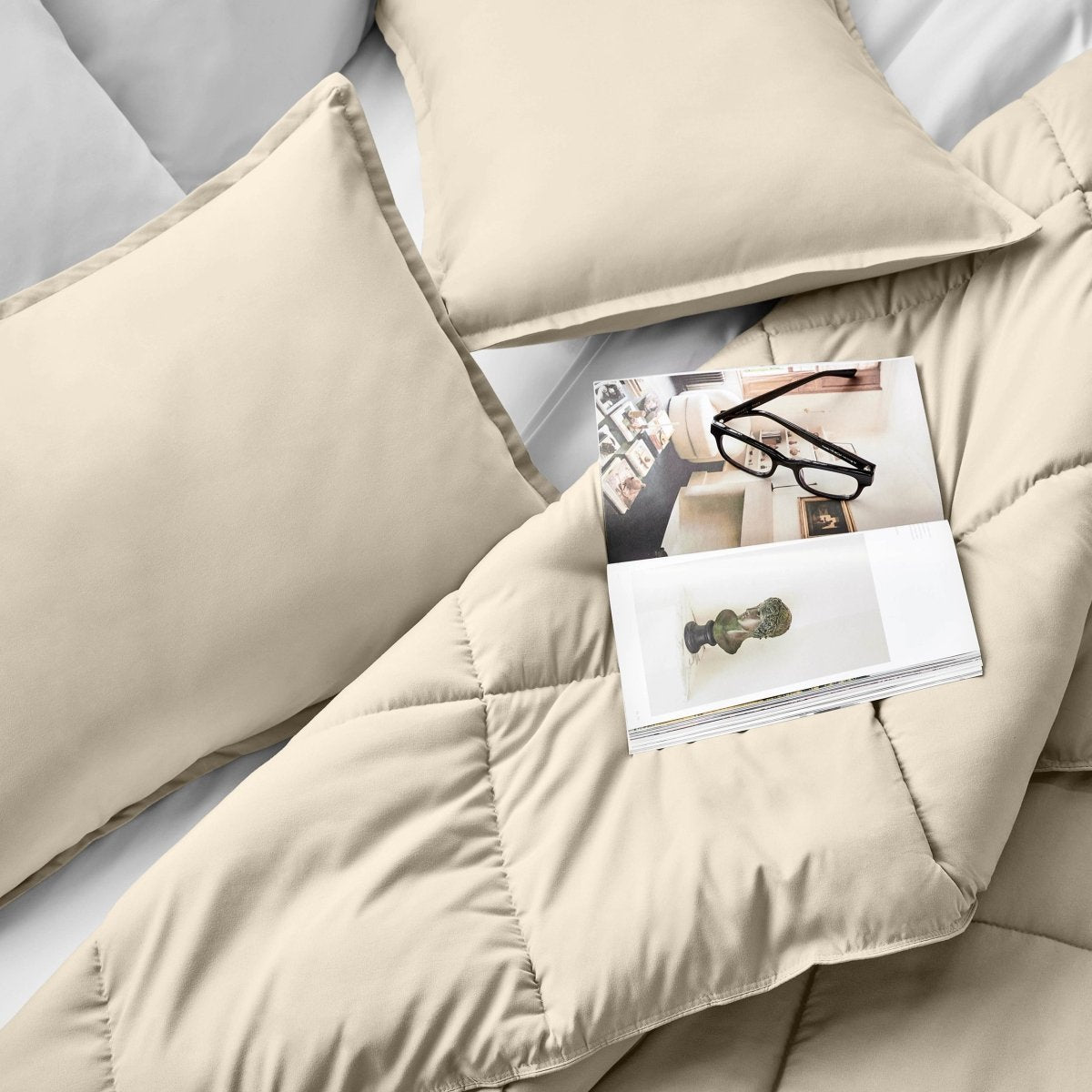 Comforter and pillows on a bed with a magazine and glasses.
