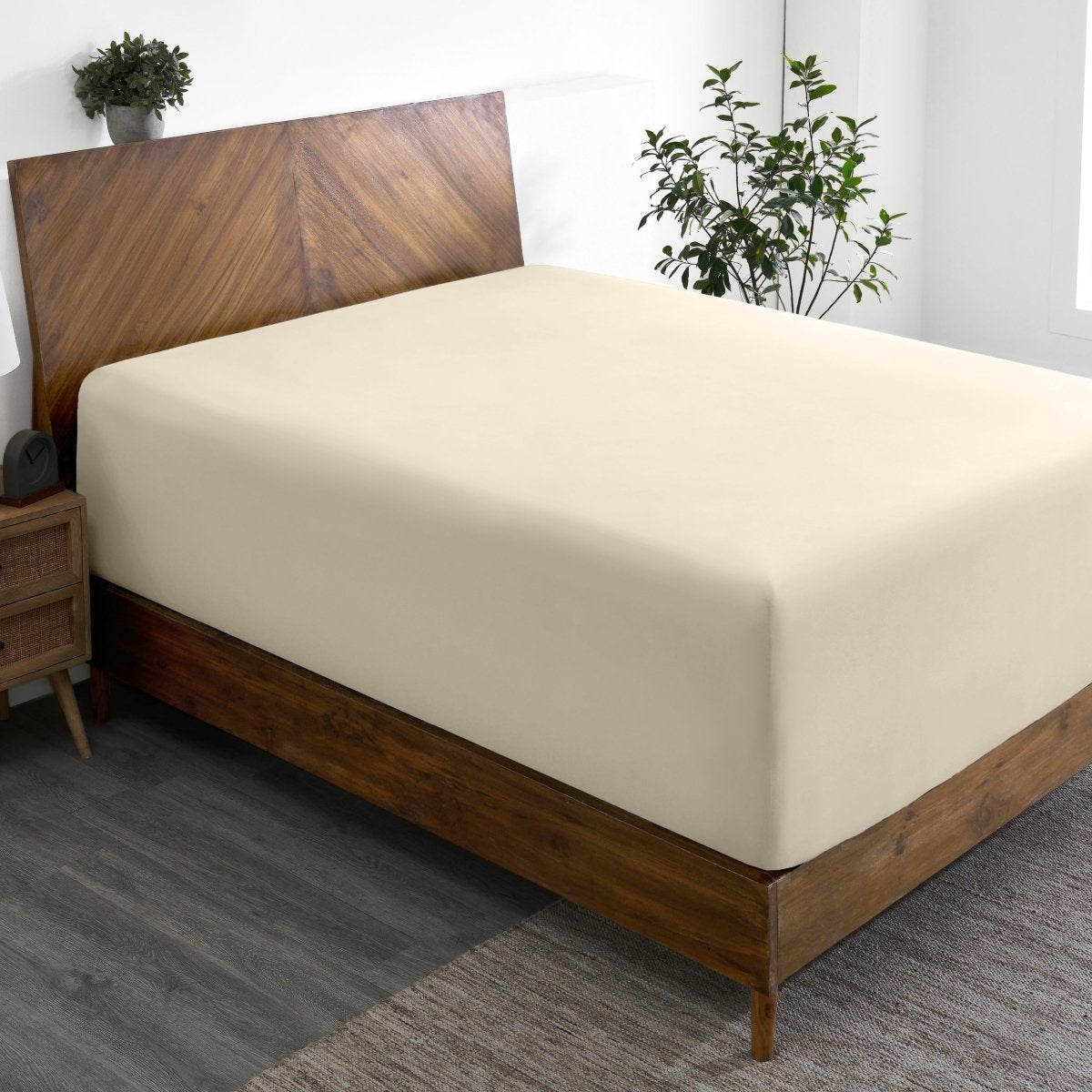 Wooden bed frame with a fitted sheet on the mattress in a room with plants and a nightstand.